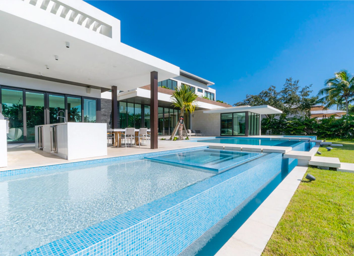 Modern luxury estate backyard featuring a blue tiled swimming pool, integrated jacuzzi, outdoor kitchen, and patio.